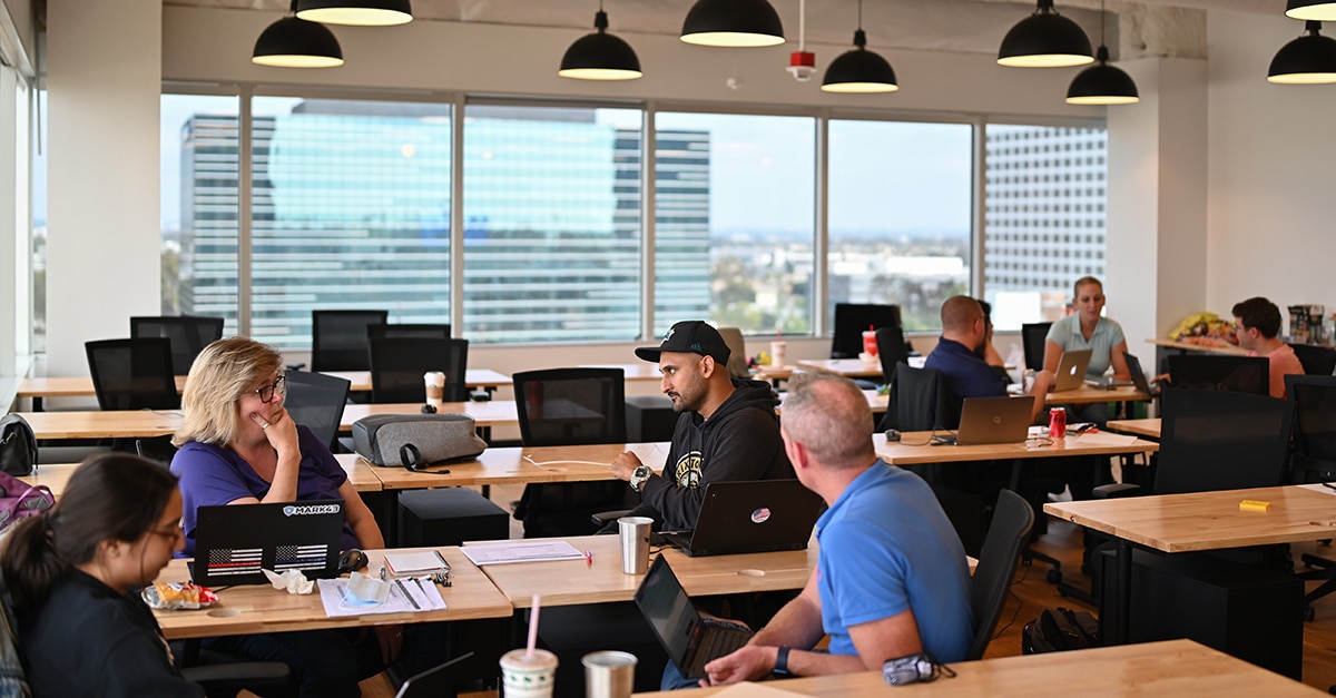 In the foreground, four people sit around a table discussing hackathon ideas. In the background, there’s another group of four people sitting around a different table discussing hackathon ideas.