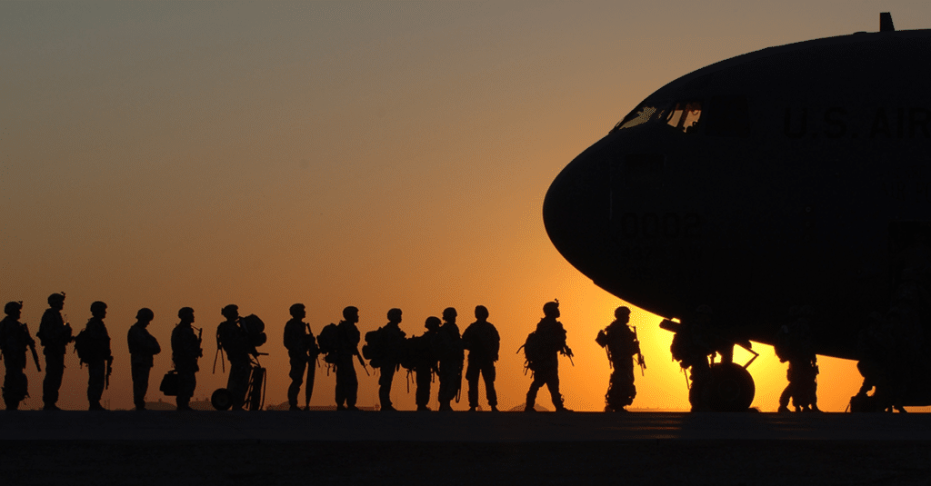 In front of a deep orange sunset, silhouettes of armed forces members wait in a line to board a large military aircraft