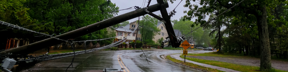 A tilted utility pole and downed power lines blocking a wet road in a residential neighborhood on a rainy day.