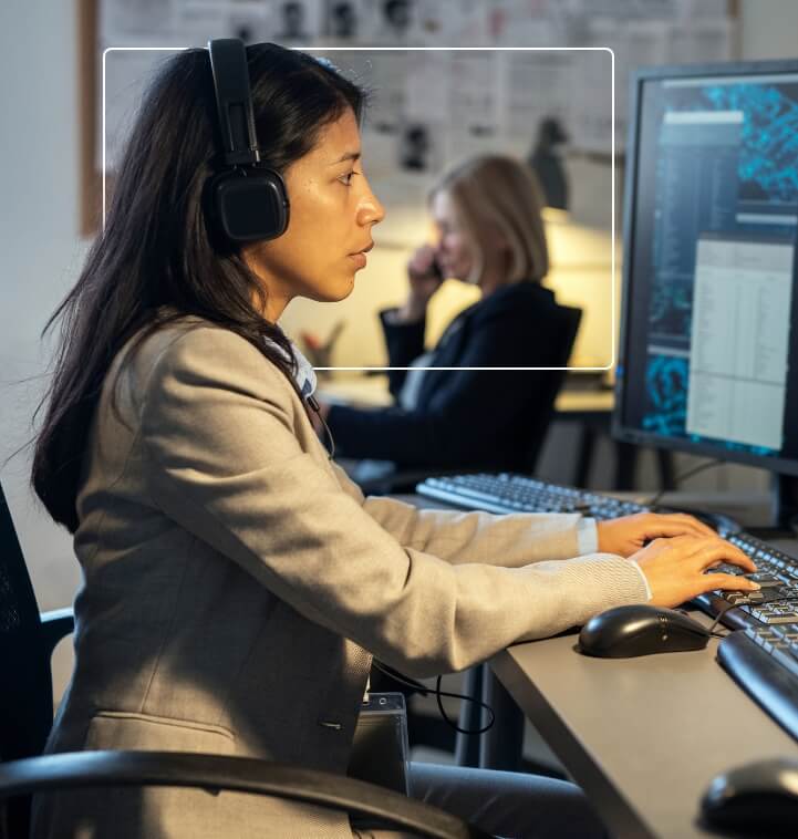 Focused woman in a headset working at a computer with a colleague in the background on the phone