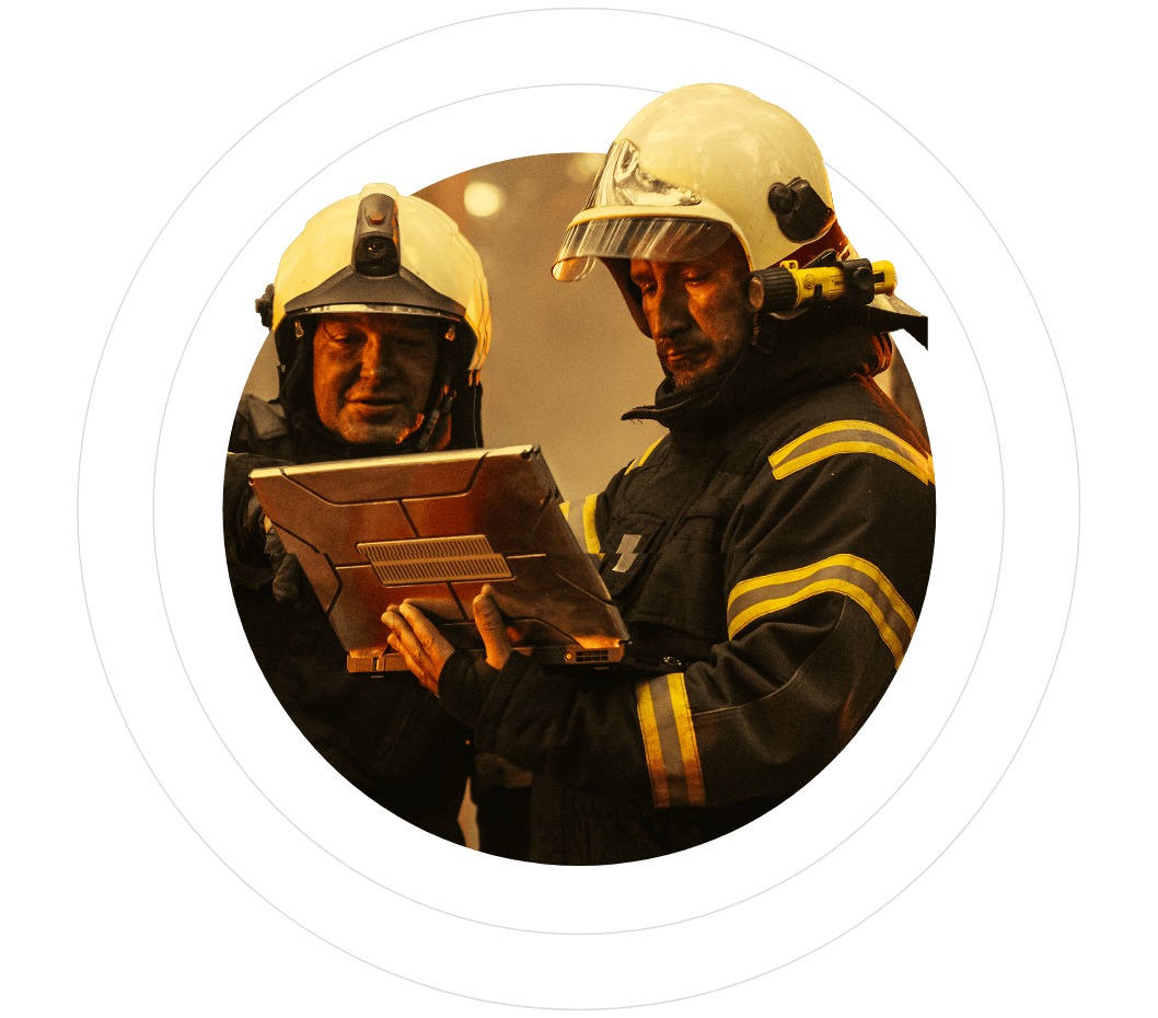 Two firefighters using a tablet, wearing helmets and protective gear