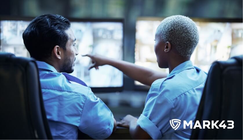 Two uniformed officers are monitoring surveillance footage on multiple screens, with one officer pointing at a screen.