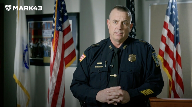 Police officer in front of American flags, speaking to the camera