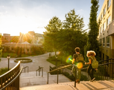 Two students with backpacks walking up steps on a sunny campus with trees and buildings in the background.