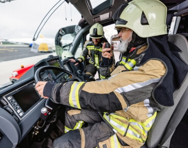 Firefighters in a vehicle, one speaking into a radio, wearing full protective gear and helmets