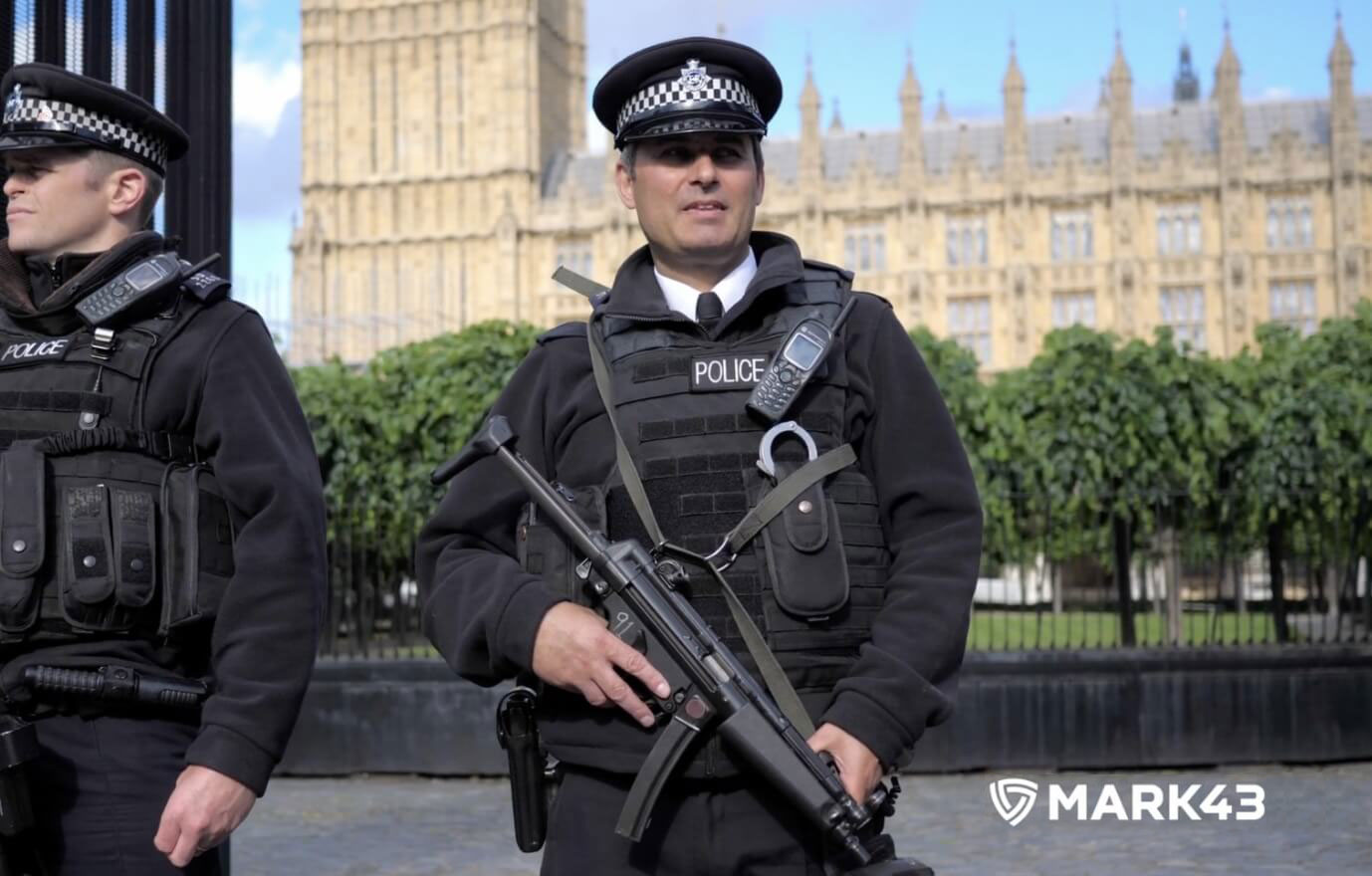 Two police officers in tactical gear, one holding a rifle, standing in front of a historic building.